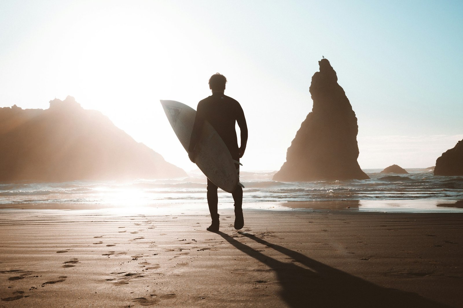 Surfer walking along the beach with a surfboard, backlit by the sun